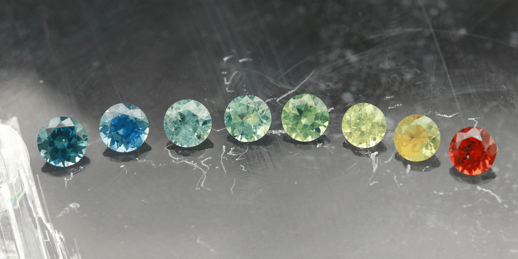 An array of eight faceted round American sapphires in a rainbow of colors. From left to right: dark teal, true medium blue, light aqua teal, sea foam green, medium green, light yellowish green, yellow, deep red orange.