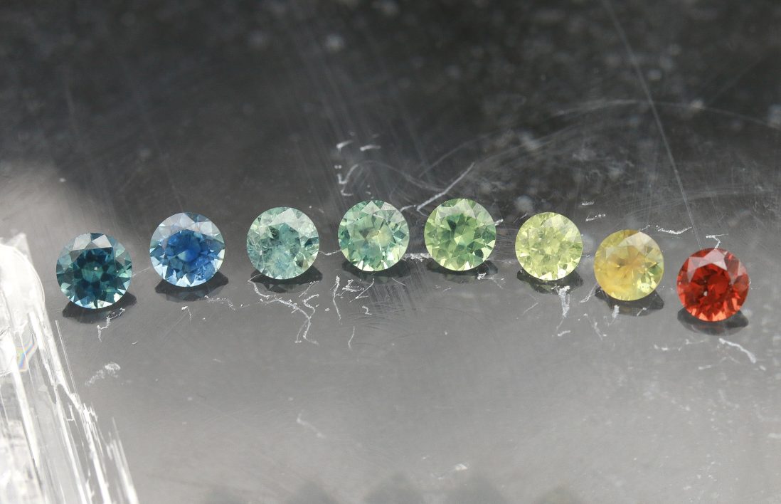 An array of eight faceted round American sapphires in a rainbow of colors. From left to right: dark teal, true medium blue, light aqua teal, sea foam green, medium green, light yellowish green, yellow, deep red orange